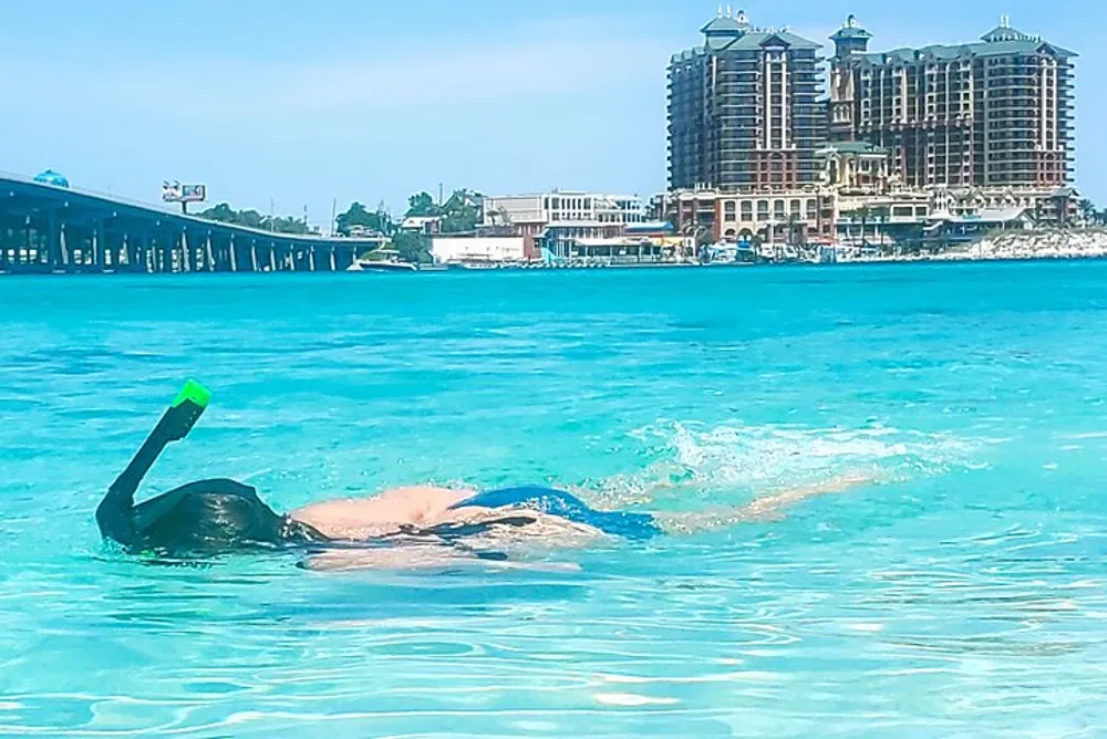A person is snorkeling in clear blue water near a pier with buildings in the background