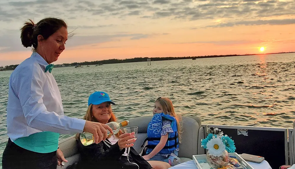 A server is pouring a drink for a smiling woman on a boat during a sunset while a young girl watches