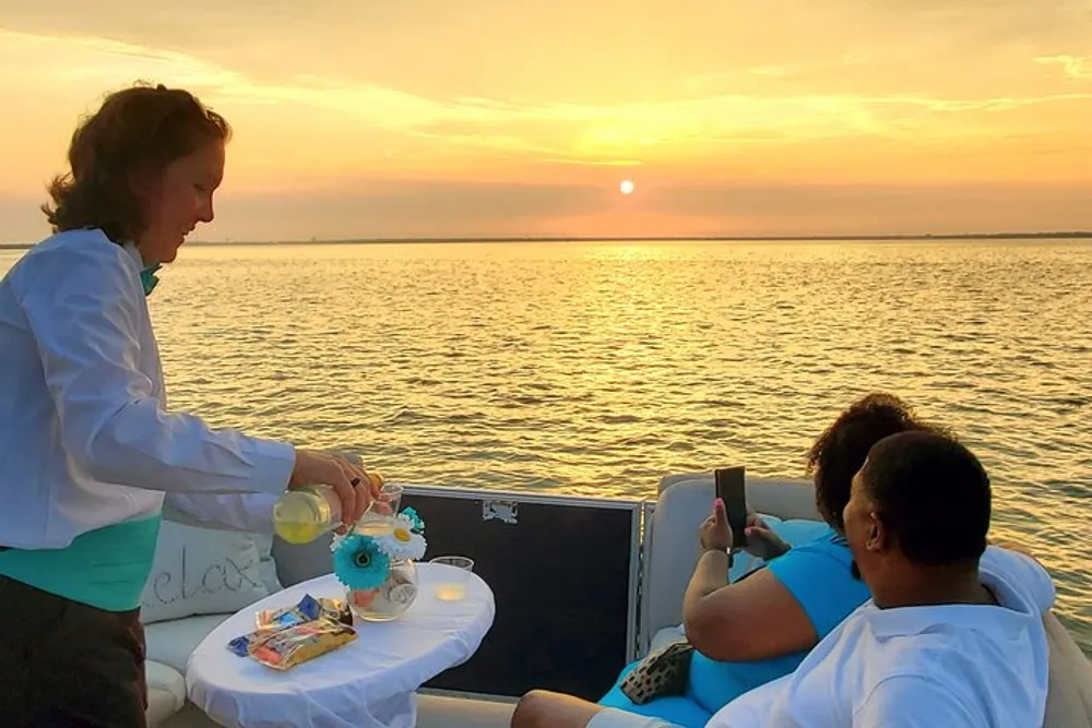 A person serves refreshments to two individuals who are enjoying a sunset on a boat