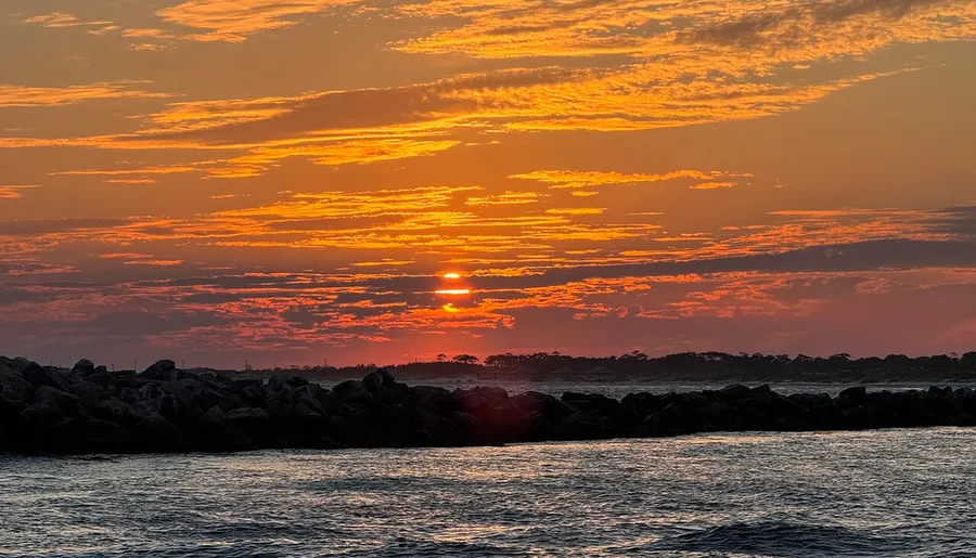 The image captures a tranquil sunset with vibrant orange hues reflecting off the clouds above a rocky shoreline and calm sea.