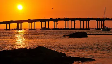 The image captures a serene sunset over the water, silhouetting a bridge structure with boats nearby, and highlighting the water with a golden glow.