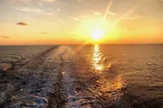 The image depicts a sunset over the ocean with the sun casting a warm glow on the water, accompanied by the gentle wake of a boat.