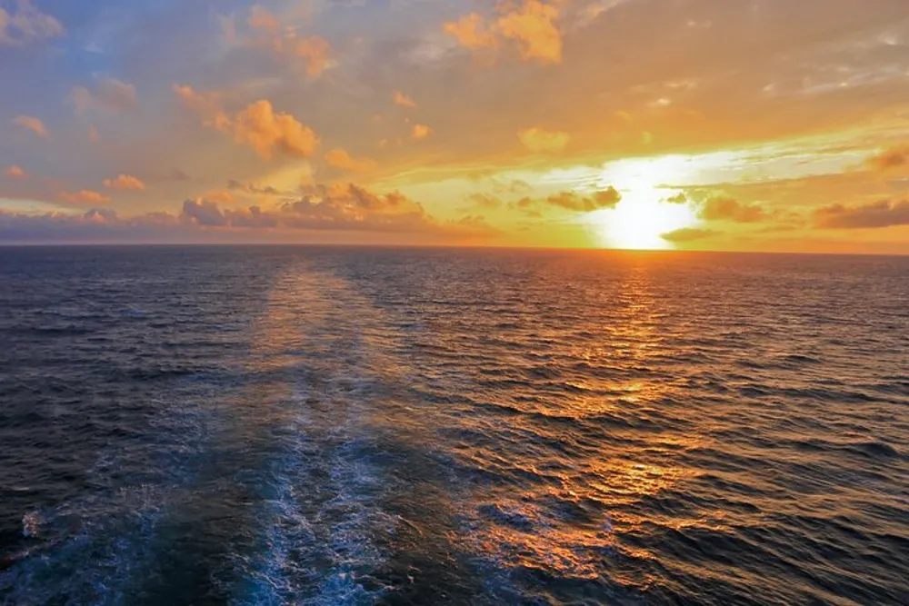 A vibrant sunset casts golden hues over the ocean with a trail of ships wake leading towards the horizon