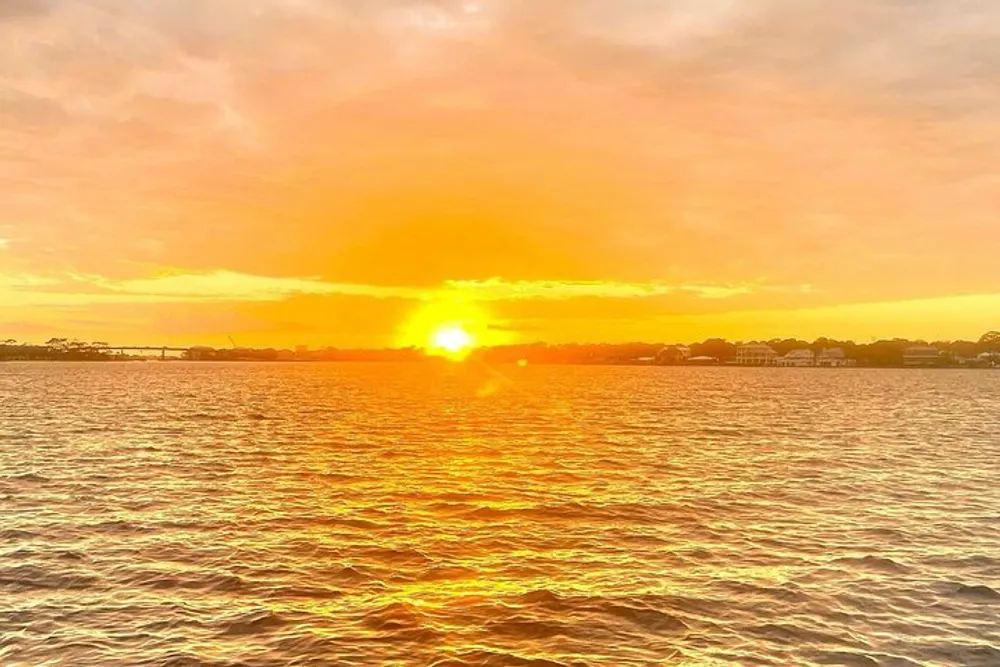 The image depicts a vibrant sunset with the sun dipping close to the horizon casting a warm golden glow over the rippling water with silhouettes of land and structures in the distance