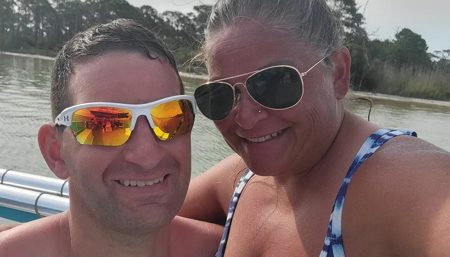 Two people are smiling for a selfie with sunglasses on, likely enjoying a sunny day on a boat with a backdrop of water and greenery.