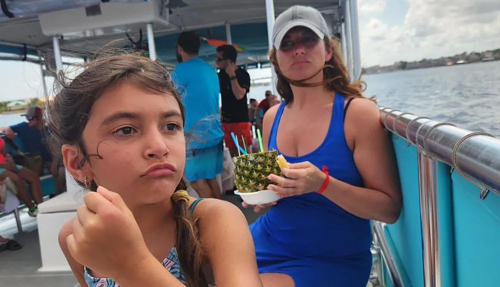 A young girl appears pensive while a woman holding a pineapple drink looks on both on a boat with other passengers in the background