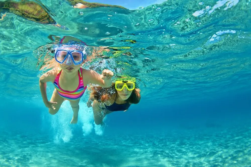 Two individuals are swimming underwater both wearing goggles with clear blue water surrounding them