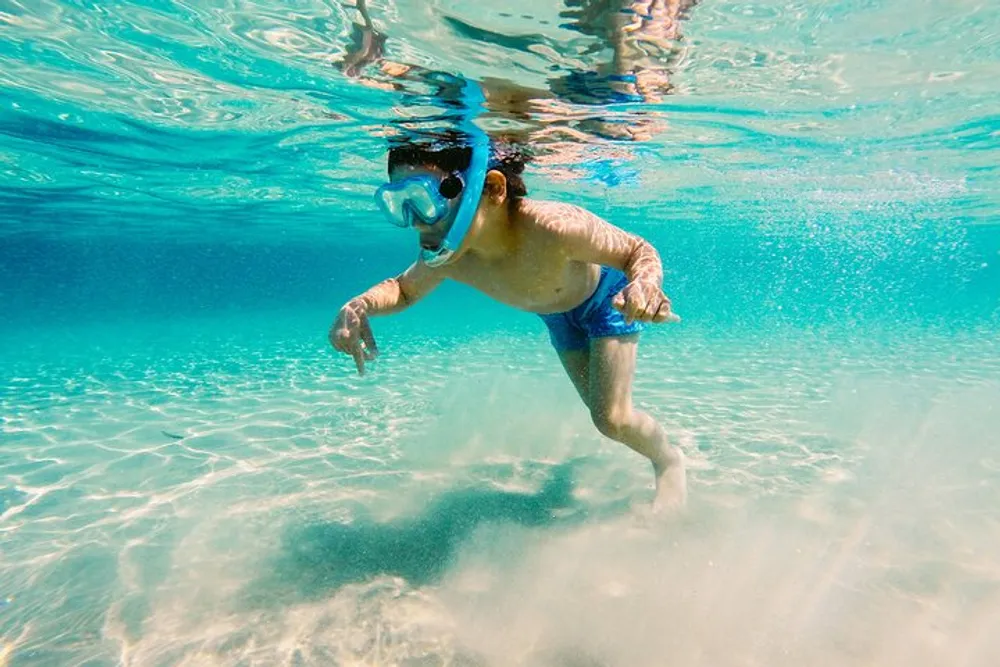 A person wearing a snorkeling mask is swimming underwater in a clear blue sea with sunlight filtering through the water