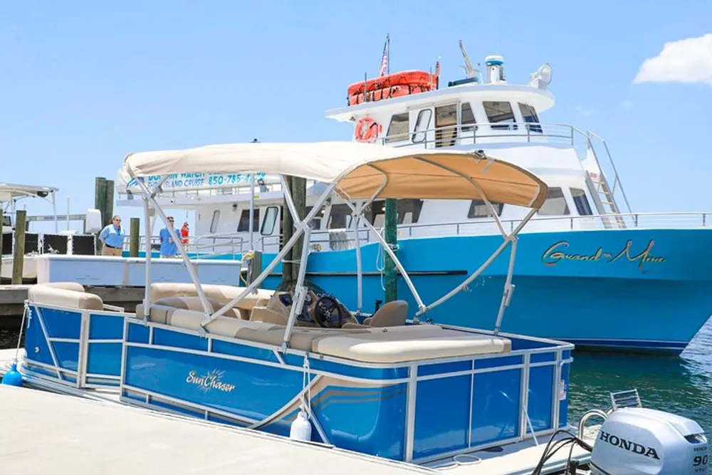A blue and white pontoon boat named Emil Chaser is docked at a marina beside a larger vessel with clear skies in the background
