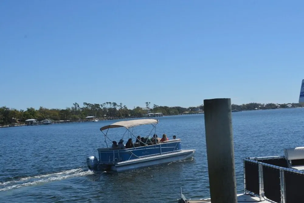 A pontoon boat with a group of people on board is cruising on a sunny day near a dock with calm waters and a verdant shoreline in the background