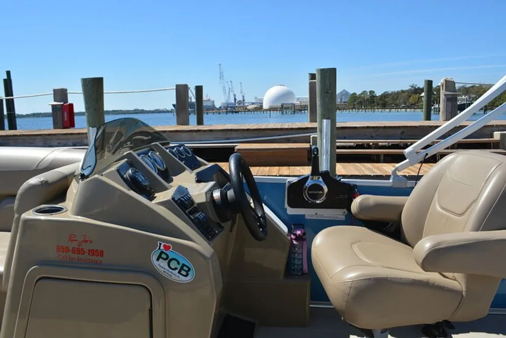 The image shows the helm of a boat with a view of a waterside featuring a dock and industrial structures in the background under a clear blue sky