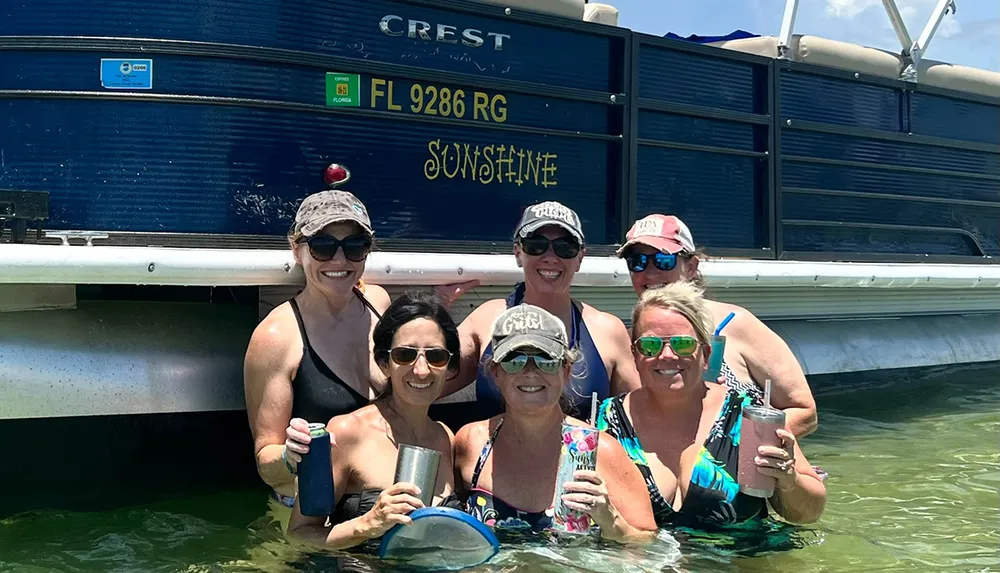 A group of six smiling people are standing waist-deep in water in front of a boat holding drinks and enjoying a sunny day