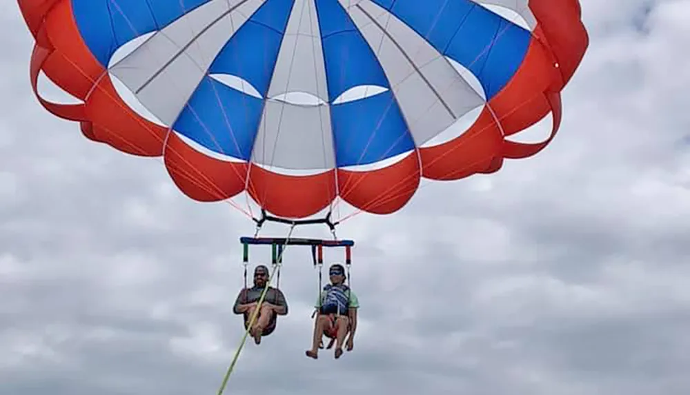 Two people are parasailing under a large red white and blue parachute against a cloudy sky