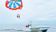 Two people are parasailing above the ocean, towed by a boat that is moving swiftly across the water.