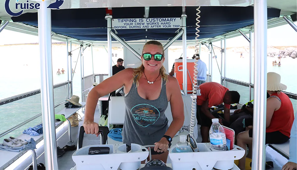 A person wearing sunglasses is smiling and standing at the helm of a boat with passengers and a sign mentioning tipping customs in the background