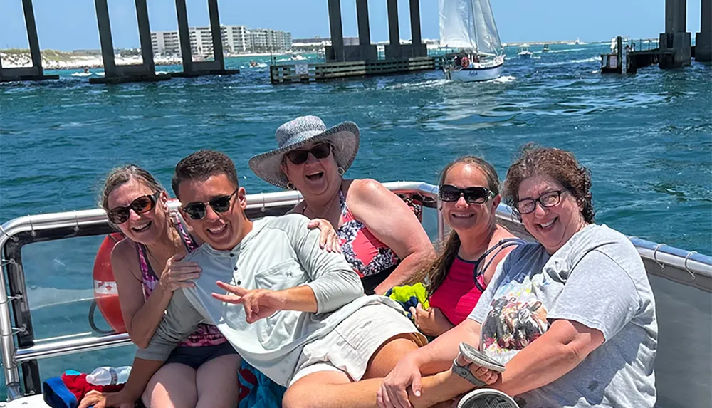 A group of people are enjoying a sunny boat ride near a bridge and a sailboat in the background smiling and posing for a photo