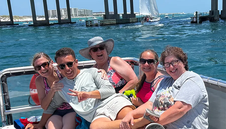 A group of people are enjoying a sunny boat ride near a bridge and a sailboat in the background, smiling and posing for a photo.