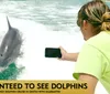 A person is photographing a dolphin leaping out of the water with a promotional text overlay guaranteeing dolphin sightings on a cruise in Destin