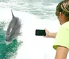 A person is photographing a dolphin leaping out of the water with a promotional text overlay guaranteeing dolphin sightings on a cruise in Destin