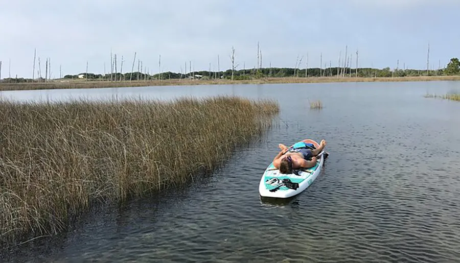 A person is lying on their back on a paddleboard in calm waters surrounded by tall grasses.