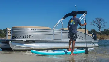 A person stands on a paddleboard near a pontoon boat on a calm water surface, suggestive of a leisurely day at the beach.