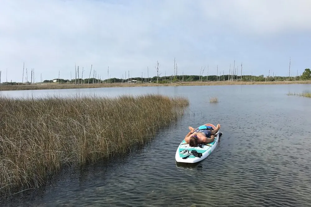 A person is lying on a paddleboard in calm water amidst tall grasses enjoying a relaxing moment