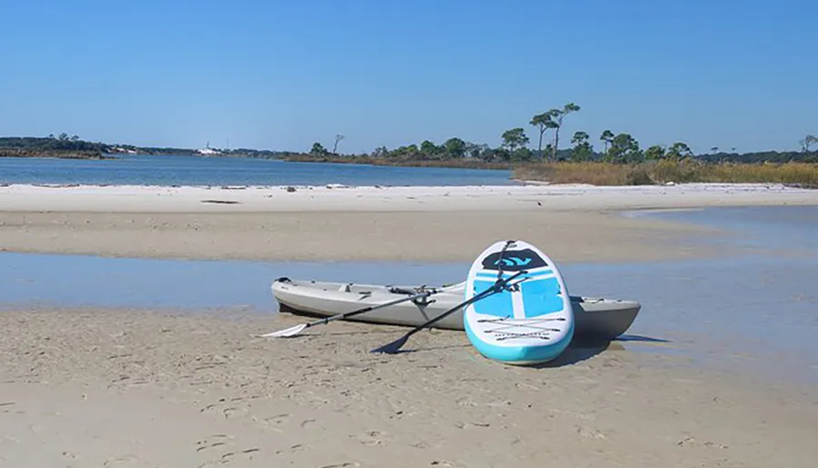 A kayak and paddleboard rest on a sandy beach near a calm body of water under a clear blue sky.