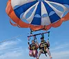 Two people are wearing life jackets and safety harnesses smiling and waving while enjoying a parasailing adventure over the water