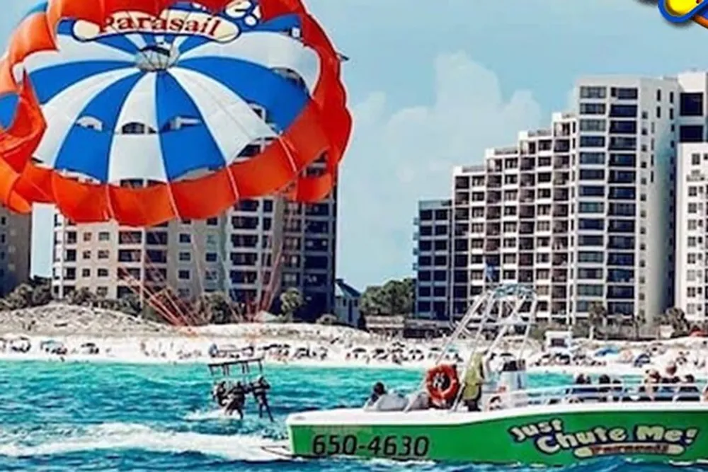 A parasailer is flying above the sea near a beachfront towed by a boat that has the slogan Just Chute Me painted on the side