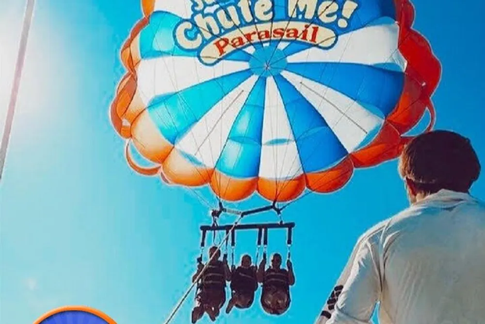 Three people are parasailing under a colorful parachute that reads Chute Me Parasail against a clear blue sky observed by another person from below