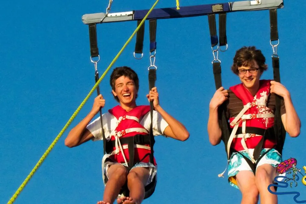 Two people are wearing life jackets and smiling while sitting on a parasailing seat against a clear blue sky