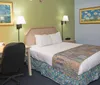 The image depicts a modestly furnished hotel room featuring a neatly made bed a desk with an office chair and decorative paintings on the wall