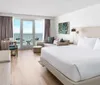 The image shows a modern hotel room with a large bed a seating area and a balcony overlooking the ocean