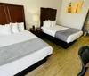 The image shows a hotel room with one large bed and one single bed both neatly made with a work desk and chair a piece of wall art and a glimpse of the bathroom