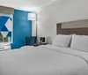 The image shows a neatly arranged hotel room with a prominent blue accent wall a large bed with white linens and a window with a geometric patterned shade