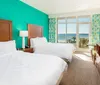 A bright hotel room featuring two beds with a turquoise accent wall leading to a balcony with an ocean view