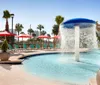 The image shows a sunny outdoor swimming pool area with a large blue and white fountain feature surrounded by red umbrellas and lounge chairs evoking a relaxing resort atmosphere