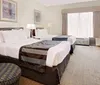 This image shows a neatly arranged hotel room with two queen-sized beds patterned chairs and a flat-screen TV projecting a comfortable accommodation space