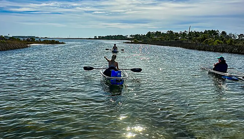 Three people are kayaking in a calm waterway flanked by vegetation under a partly cloudy sky
