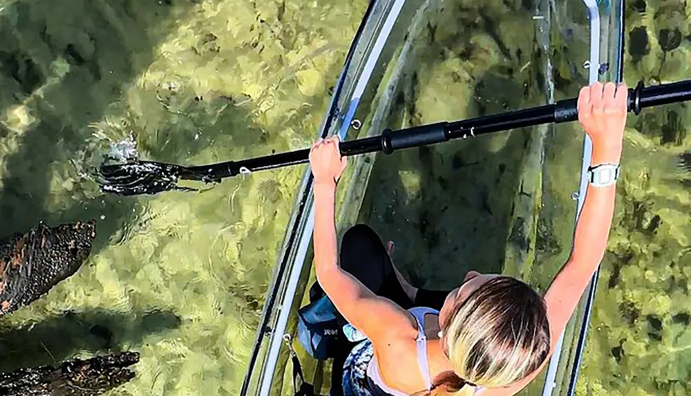 A person is paddling a transparent kayak over clear water offering a view of the underwater environment