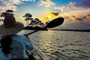 A person in a straw hat is kayaking on a calm water body during a beautiful sunset.