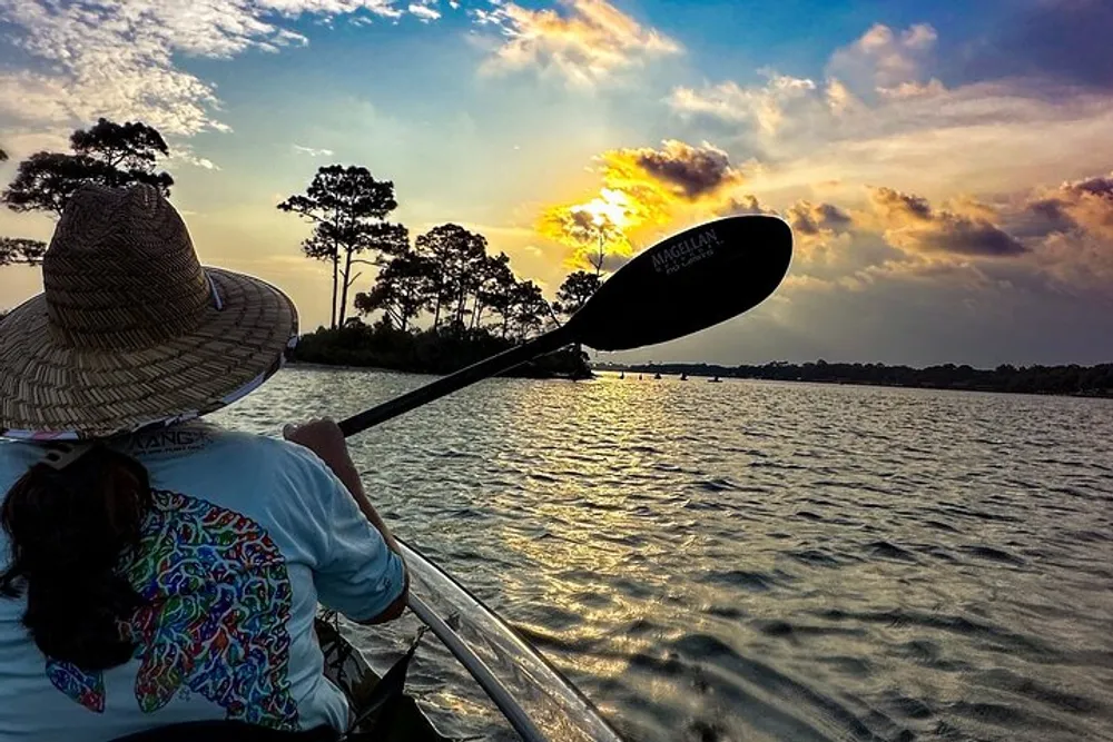 A person in a straw hat is kayaking on a calm water body during a beautiful sunset