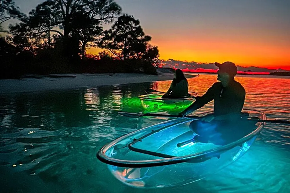 Two people in illuminated kayaks are on a serene body of water under a vibrant sunset sky