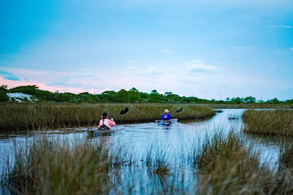 Two people are kayaking through a tranquil waterway surrounded by tall grasses under a dusky sky