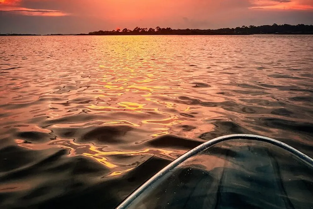 The image shows the sun setting over a rippling body of water casting warm hues of orange and red with the bow of a kayak visible in the foreground
