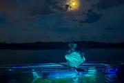 A person sits in a transparent canoe under a moonlit sky, with the water illuminated by blue light from underneath the vessel.