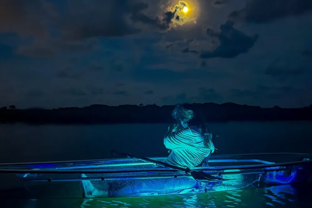 A person sits in a transparent canoe under a moonlit sky with the water illuminated by blue light from underneath the vessel