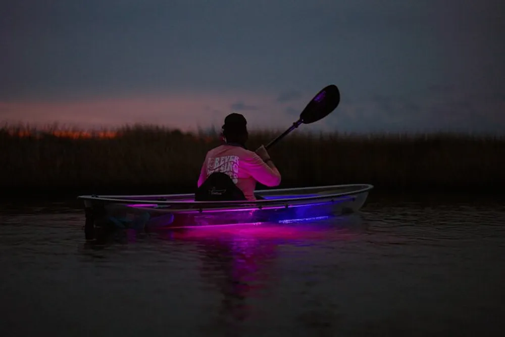 A person is paddling a translucent LED-lit kayak at dusk creating a striking contrast against the darkening sky and water