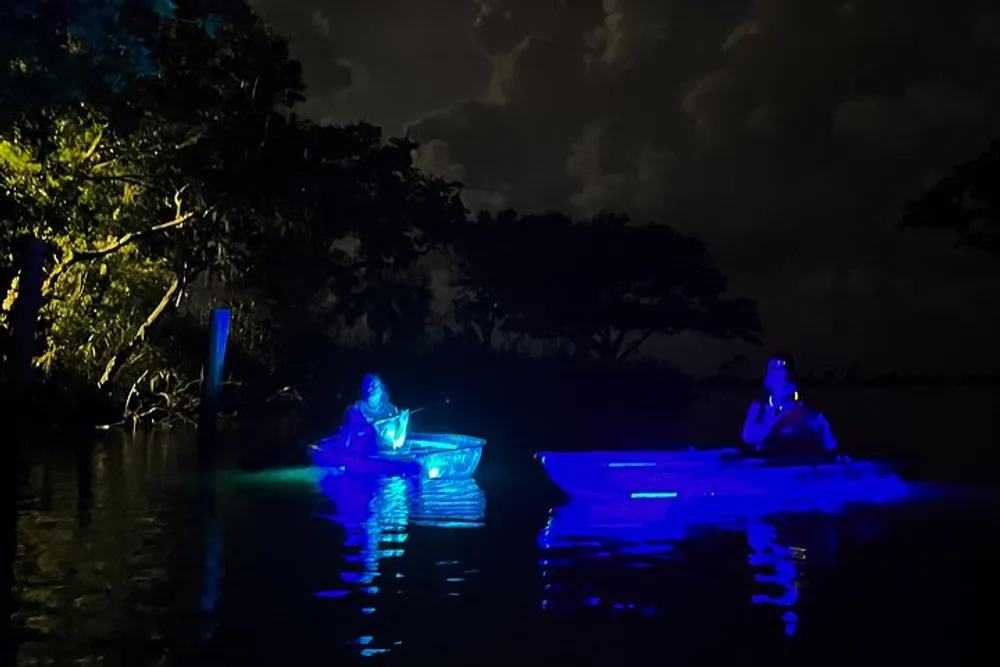 Two people are kayaking at night with their kayaks illuminated by blue lights creating an ethereal glow on the water