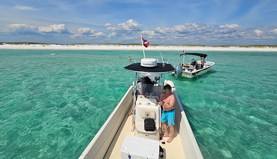 A person stands on a boat floating in clear turquoise waters near a sandy beach with other boats and people enjoying the day.